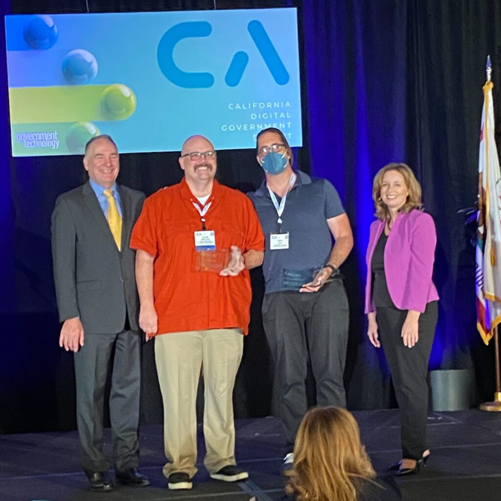 Blaine and Aaron stand onstage to receive the award, flanked by Alan and Liana. Behind them a screen displays the logo for the California Digital Summit 2022.